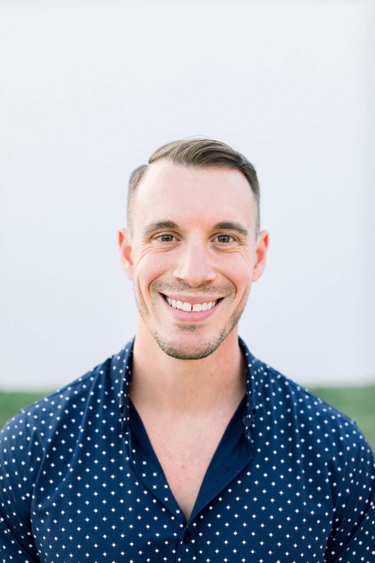 Smiling man with short hair and a trimmed beard wearing a dark blue patterned shirt against a plain background.