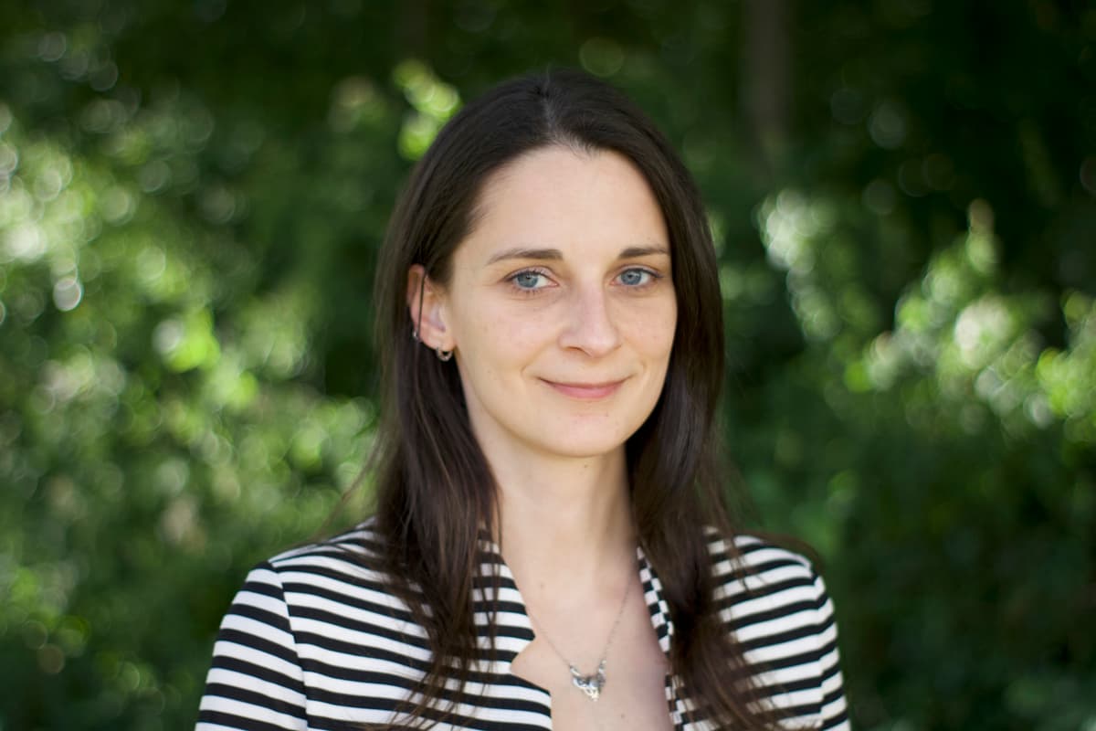 Portrait of a smiling woman with long dark hair wearing a black and white striped top against a blurred green background.
